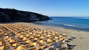Una playa con numerosos sombrillas amarillas dispuestas en filas, cielo despejado y un acantilado rocoso al fondo.