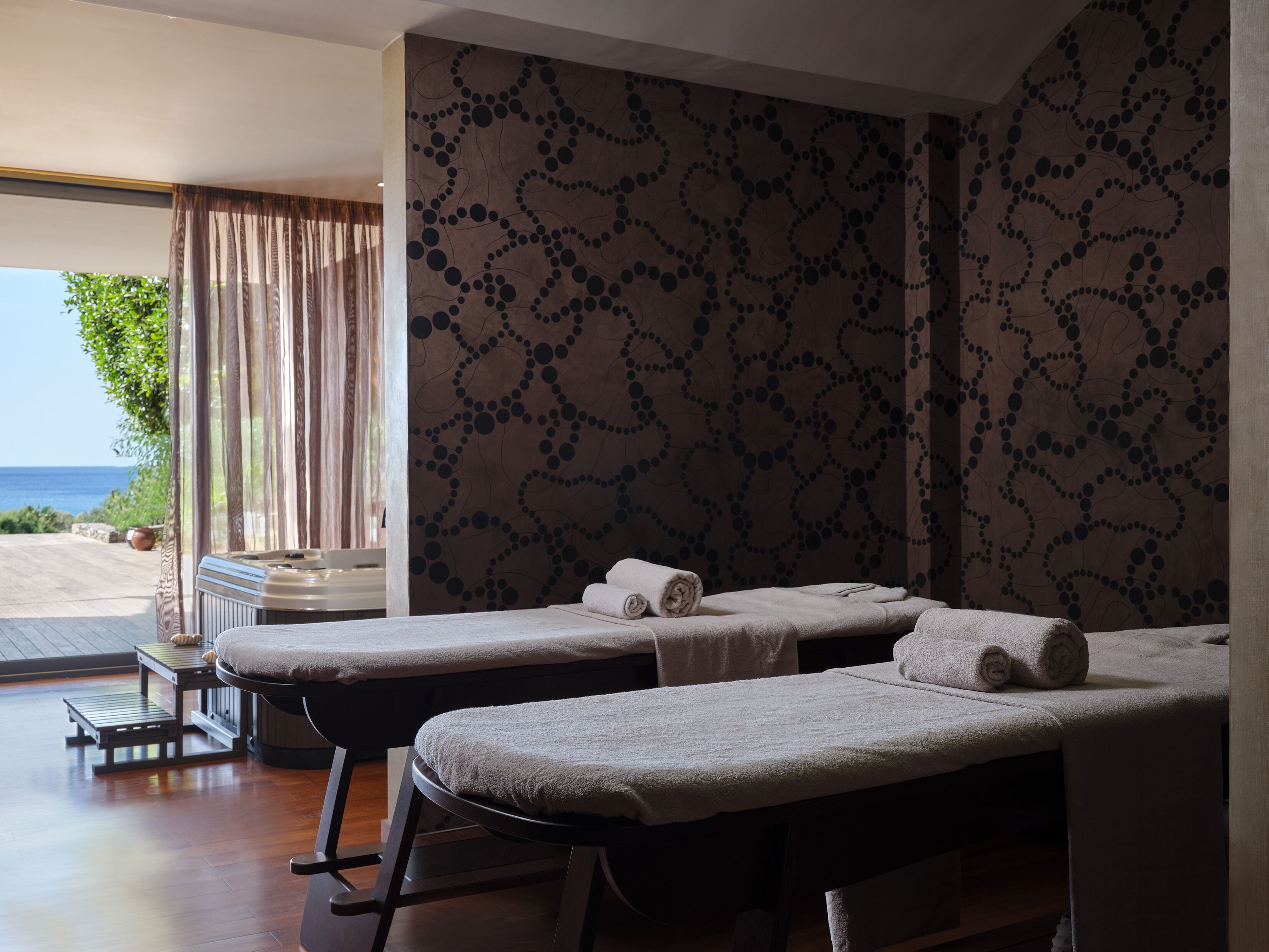 Ein Spa-Behandlungsraum mit zwei Massageliegen, Blick auf das Meer und einer Wand mit Muster.