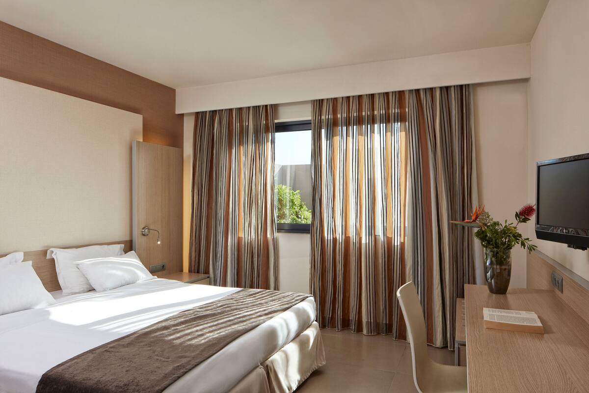 A hotel room with a large bed, a desk with a flat-screen TV, a window with curtains, and a vase with flowers.