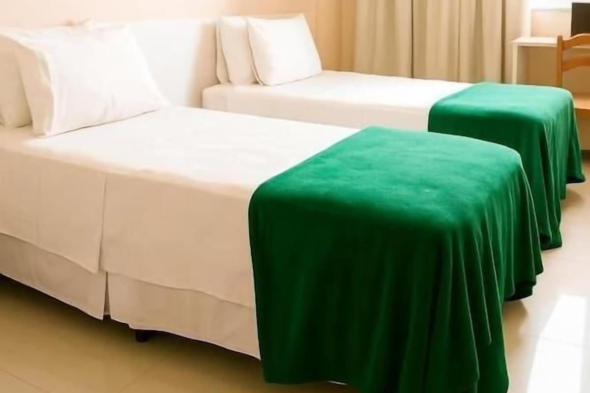 A hotel room with two beds, a desk, a chair, a window with curtains, and a framed picture on the wall.