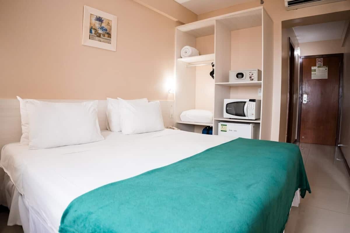 A hotel room with a bed, a green throw blanket, a microwave, and a shelf with toiletries.