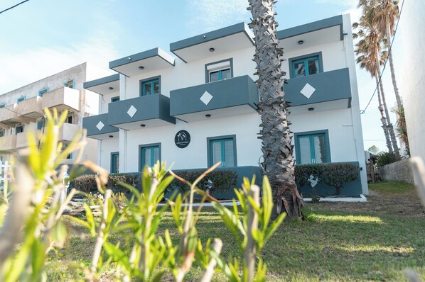 A modern building with a white facade and teal accents, featuring balconies and a small garden area. - Angela & Thalia Apartments (Kos)