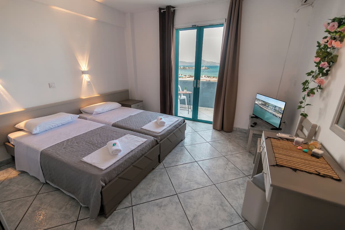 A hotel room with two beds, a TV, and a balcony with a view of the sea.
