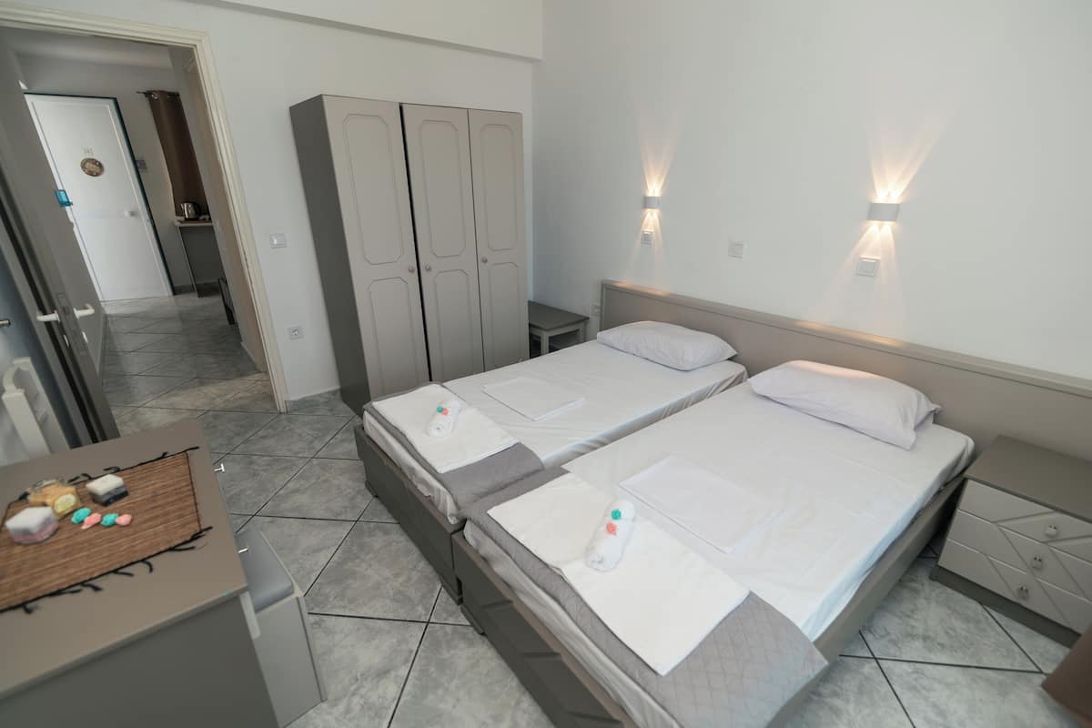 A hotel room with two beds, a wardrobe, a bedside table, and a small desk with a bottle and snacks.