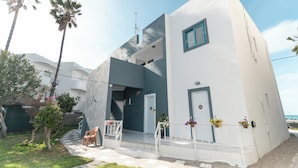 A modern two-story building with a white facade and blue accents, featuring a staircase leading to the upper level, outdoor seating, and a view of the sea. - Angela & Thalia Apartments (Kos)