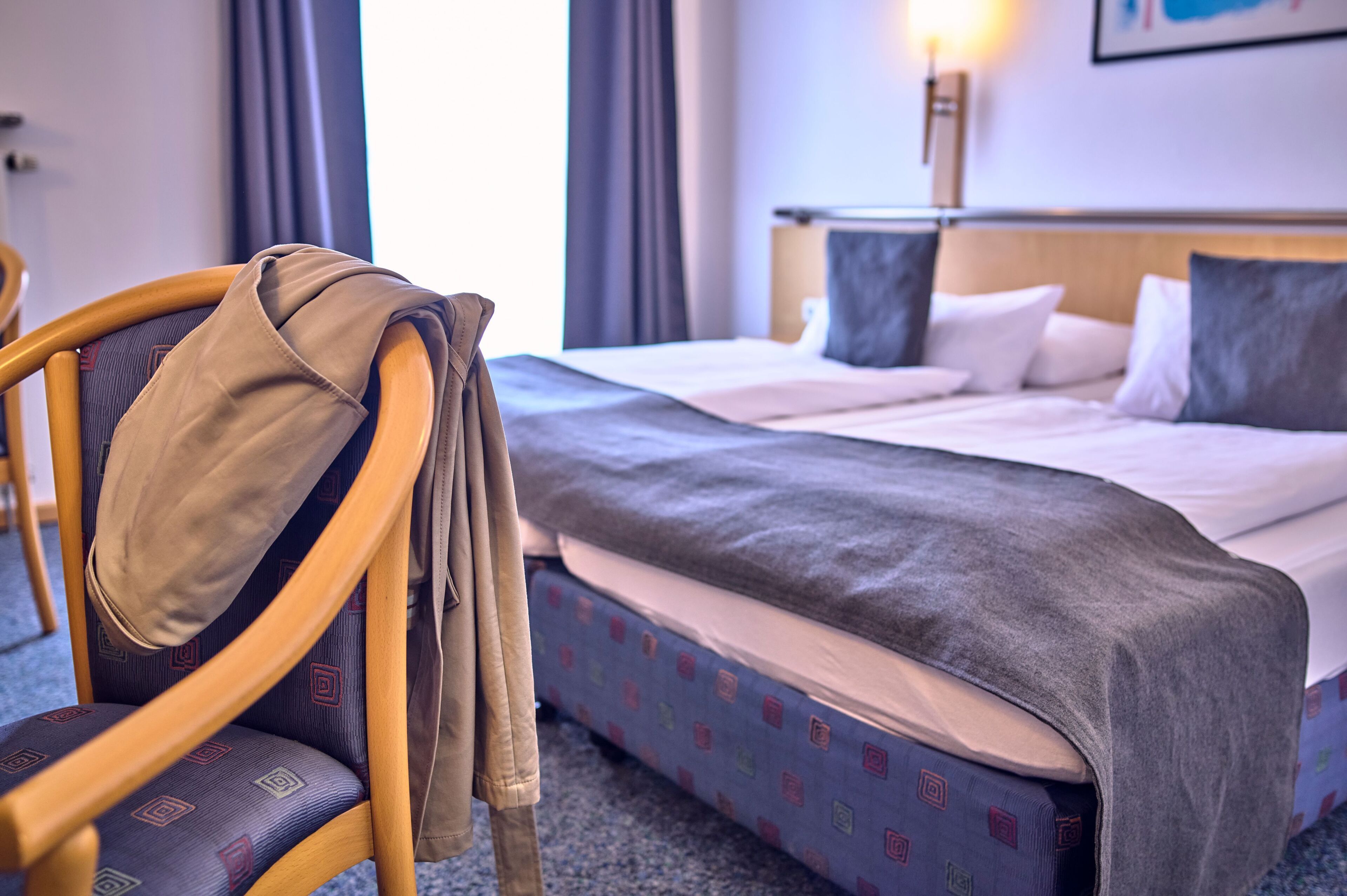 Una habitación de hotel con una cama, una silla con una chaqueta y una lámpara fijada a la pared.