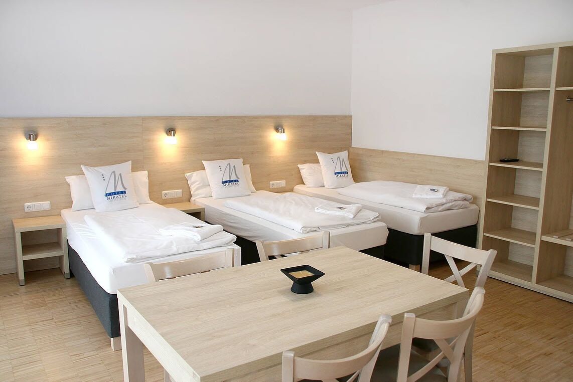 Habitación de hotel con tres camas individuales, una mesa de madera y una estantería.
