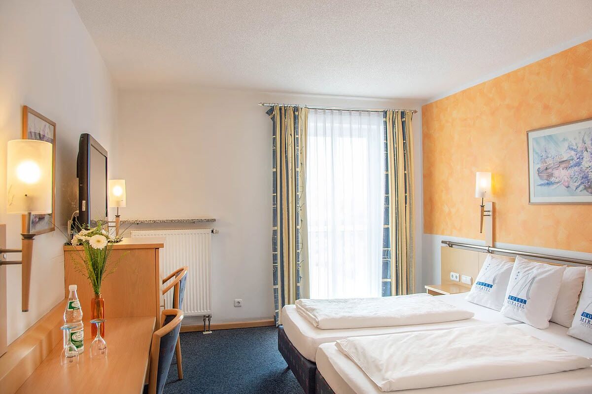 A hotel room with two beds, a desk with a chair, a radiator, and a window with curtains.