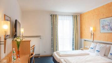 A hotel room with two beds, a desk with a chair, a radiator, and a window with curtains.