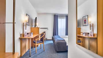 A hotel room with a bed, a desk with a chair, a small table, a radiator, a window with curtains, and a framed picture on the wall.