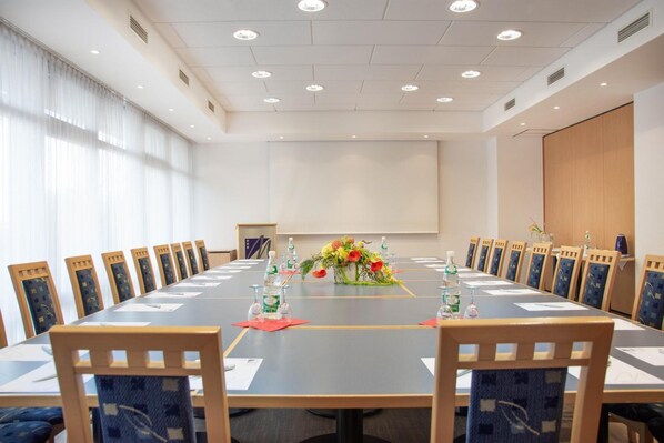 A conference room with a long table, chairs, a projector screen, and floral centerpieces. - Miratel Hotel (Mitterteich)