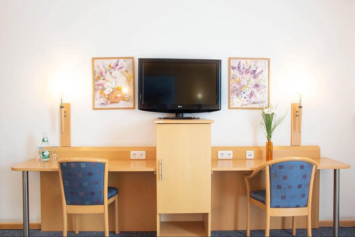 Une chambre d’hôtel avec un bureau en bois, deux chaises, une télévision et deux œuvres d’art florales encadrées accrochées au mur.