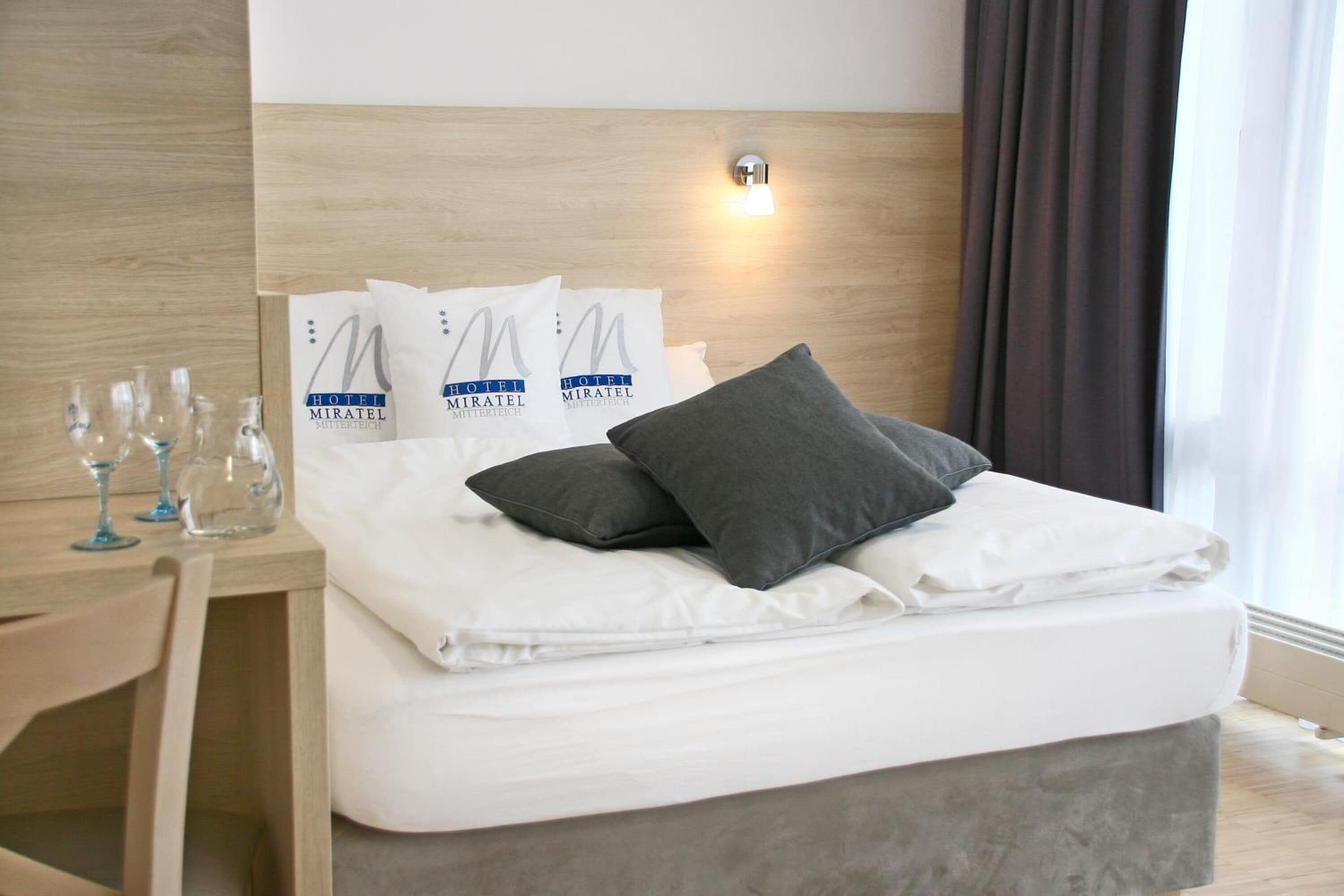 Una cama bien hecha con almohadas, una mesita de noche con vasos y una lámpara fijada a la pared.
