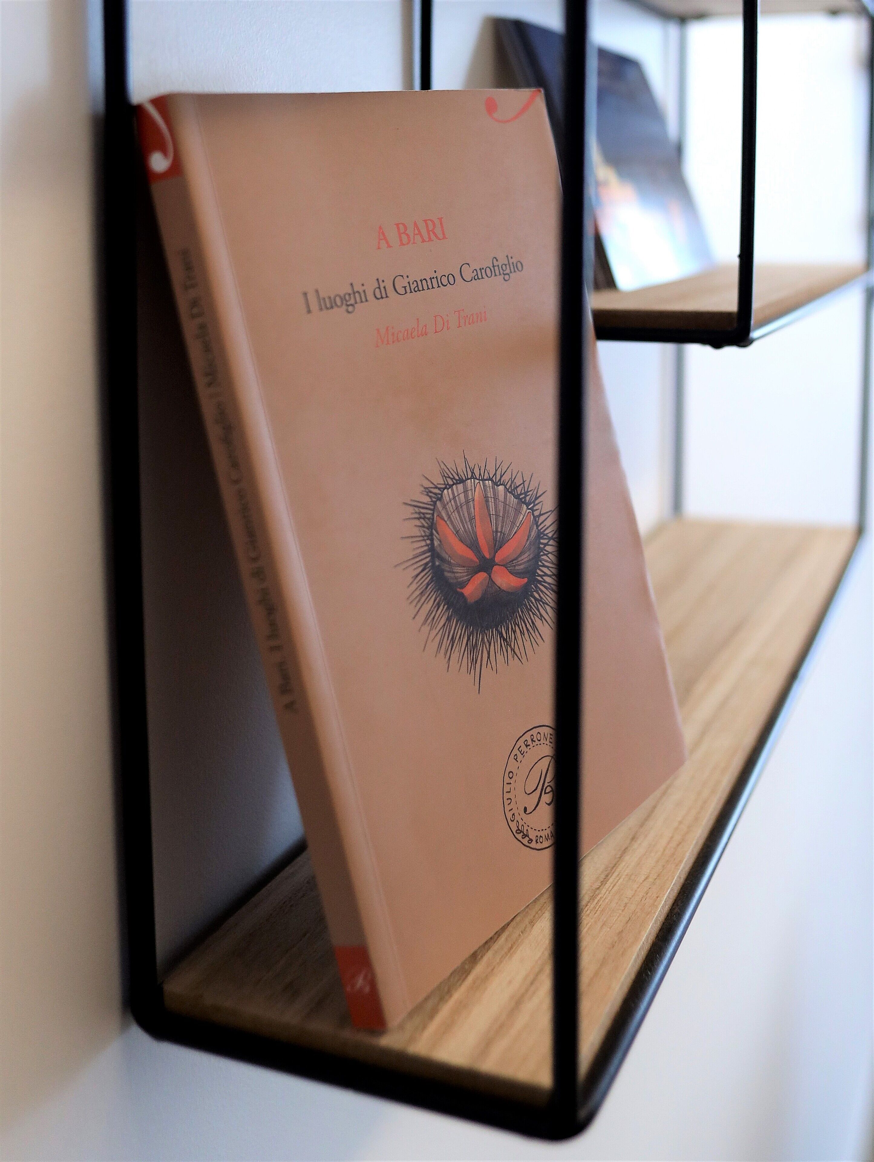 Un libro titulado "I luoghi di Gianrico Carofiglio" de Micaela Di Triani, exhibido sobre un atril de metal negro frente a una pared blanca.