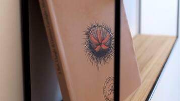 Un libro titulado "I luoghi di Gianrico Carofiglio" de Micaela Di Triani, exhibido sobre un atril de metal negro frente a una pared blanca.