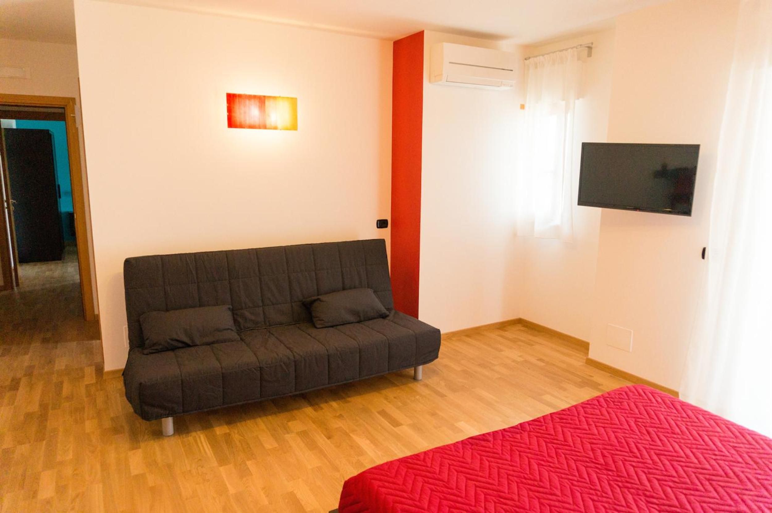 A room with a sofa, a bed with a red blanket, a television, and an air conditioning unit.