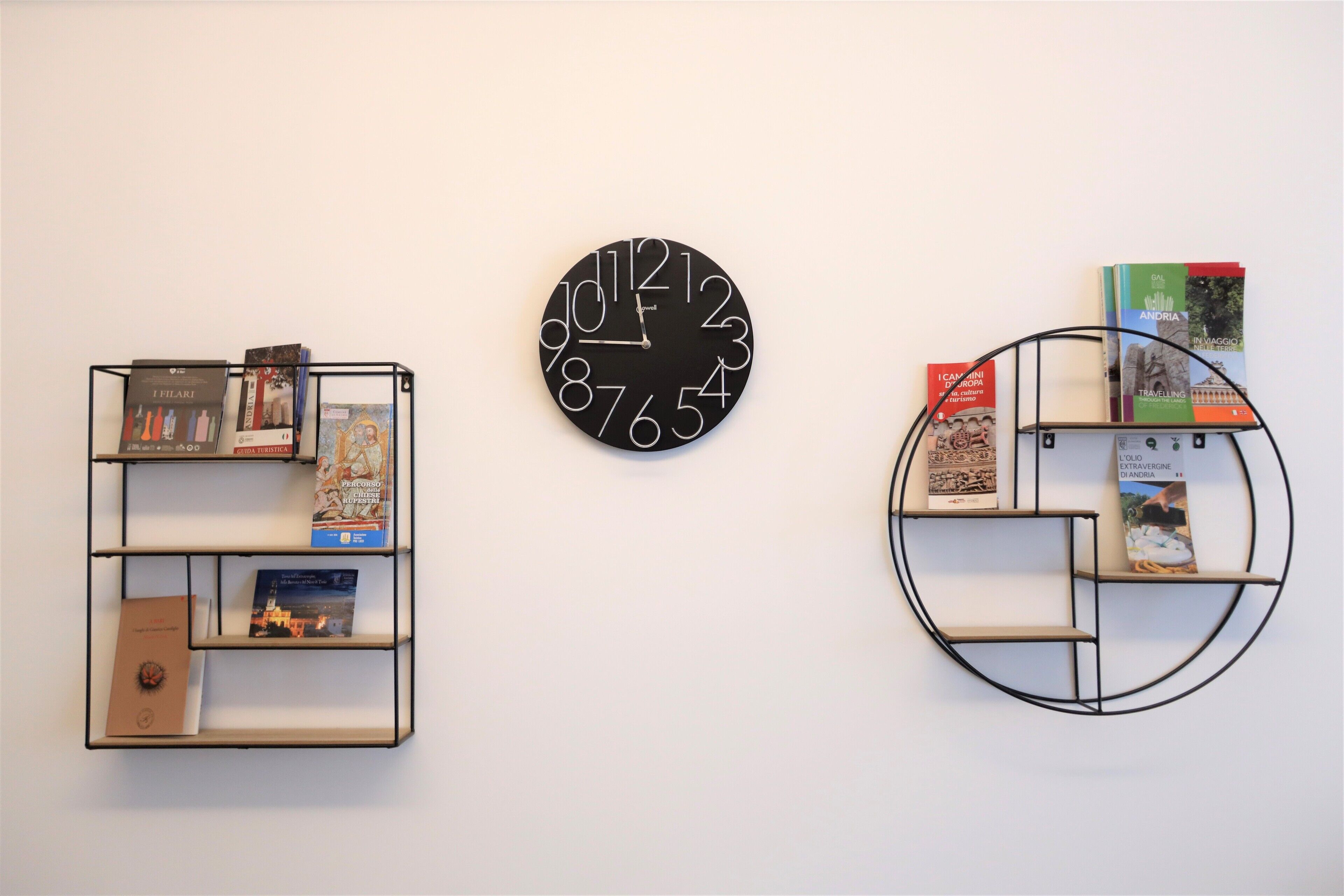 Una pared con un reloj redondo negro y dos estantes metálicos negros con libros.