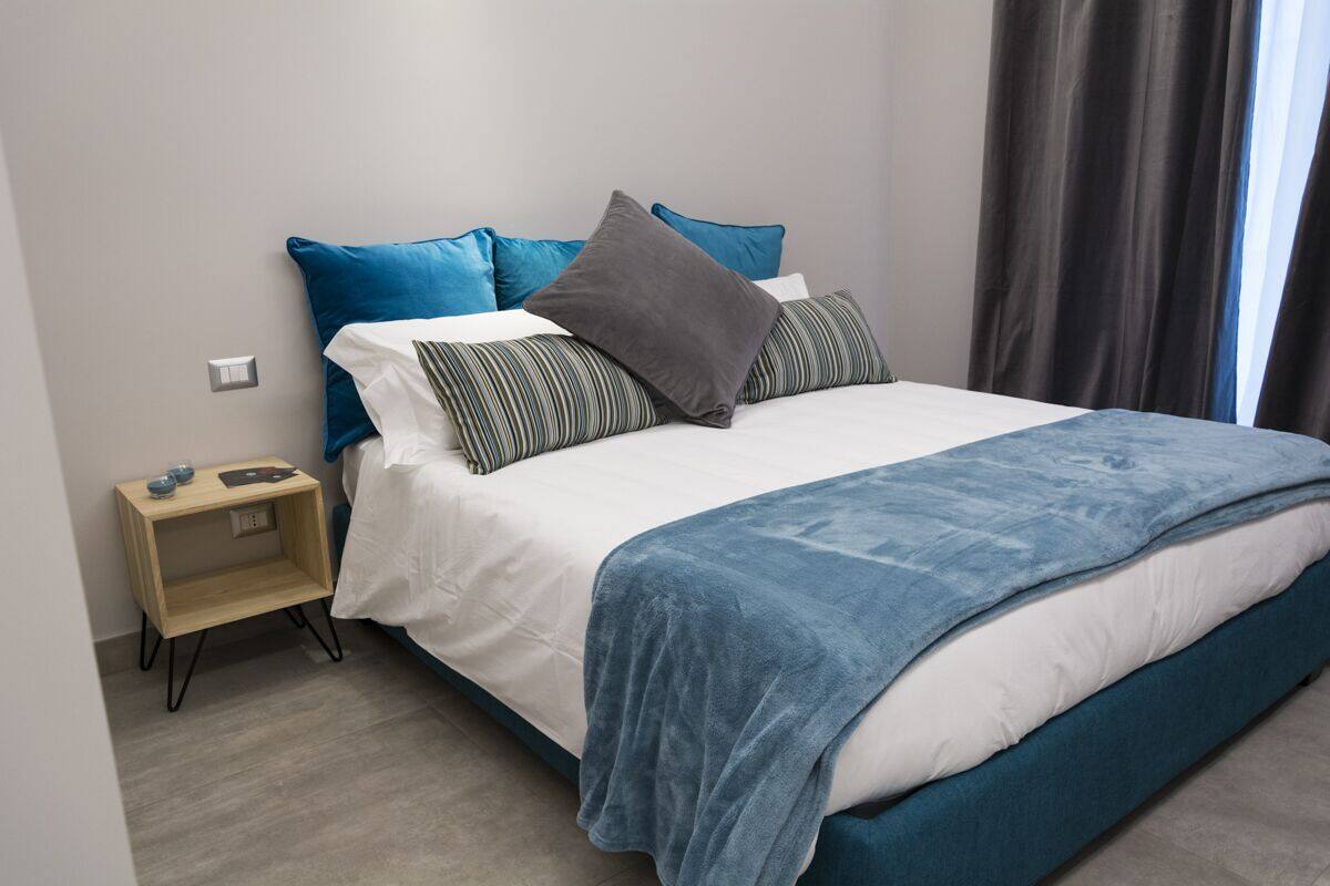 Un lit bien fait, avec une couverture bleue et des oreillers, une table de chevet avec une lampe, et une fenêtre avec des rideaux.