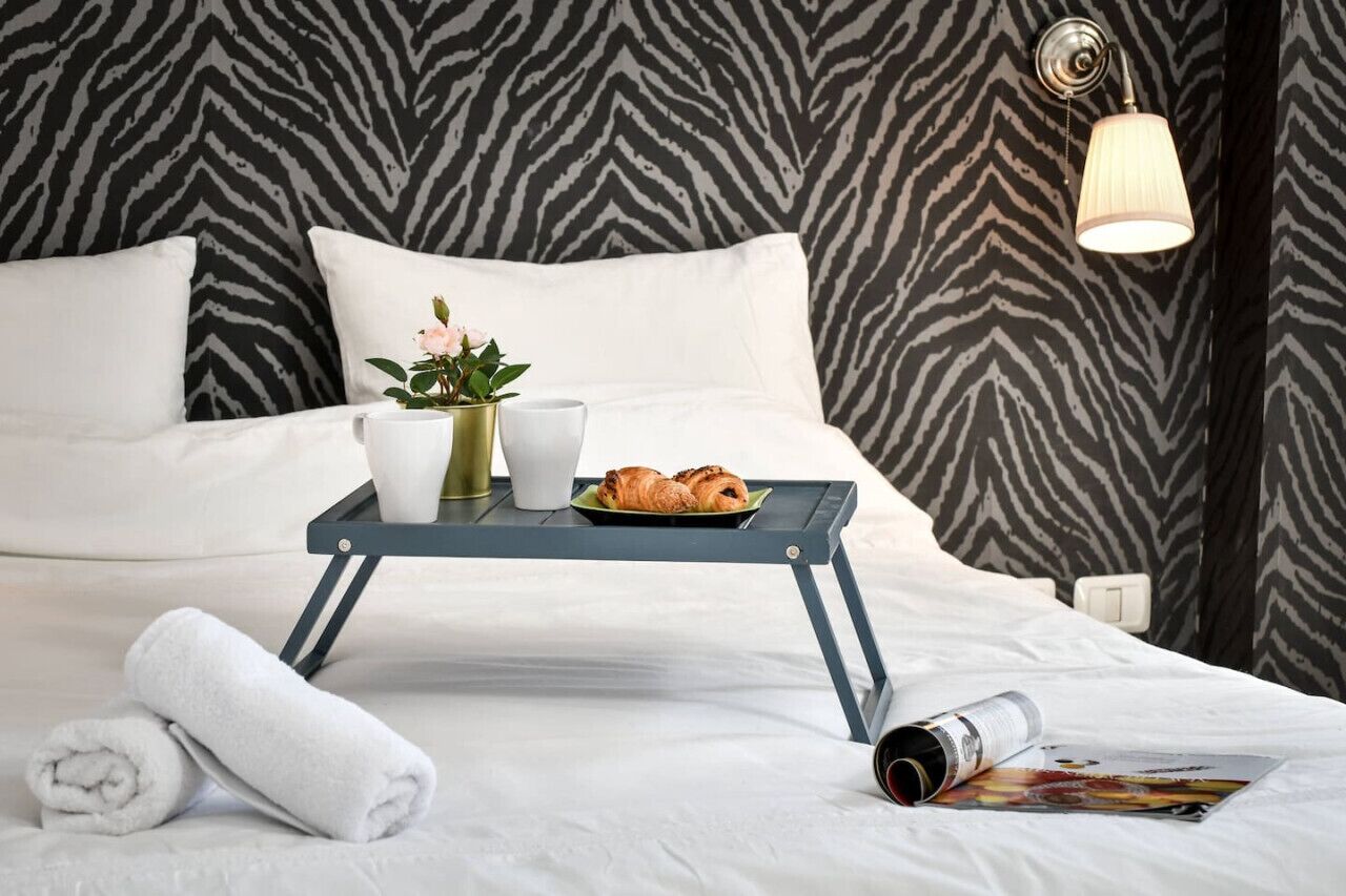 A neatly made bed with white linens, a tray with croissants and a coffee cup, a vase with flowers, and a wall with a zebra print pattern.