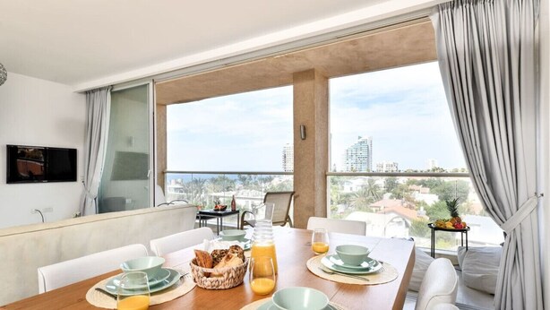 A dining area with a wooden table set for breakfast, large windows with city views, and a balcony with a view of the cityscape. - Modern Gem with Stunning Balcony by Sea N Rent (Herzliya)