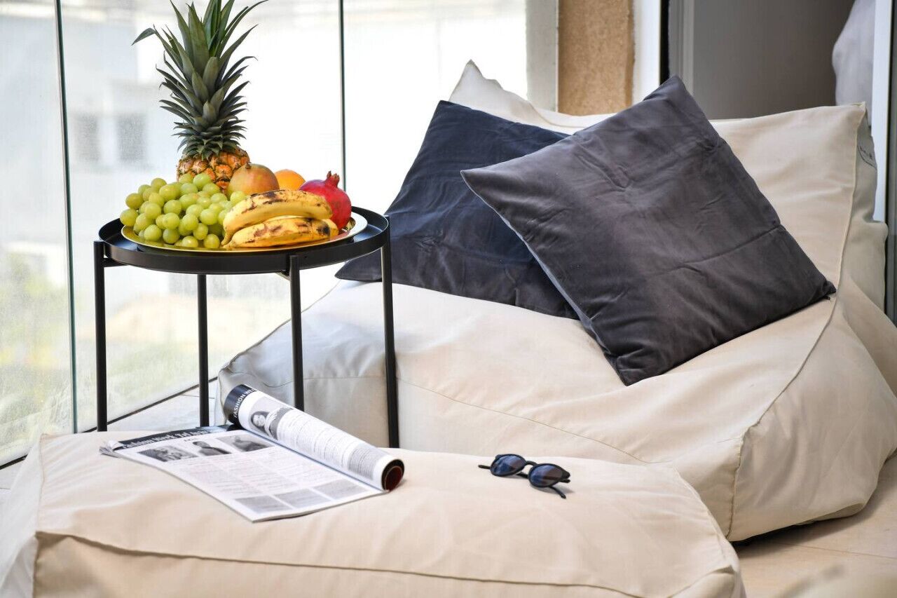 Un coin salon confortable avec un banc rembourré, une petite table sur laquelle sont disposés des fruits et un journal, et une fenêtre donnant sur un beau paysage.