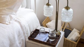 Un letto rifatto con un copriletto bianco e cuscini, un comodino con un boccale di vetro, una caraffa e un vassoio con un panno. Ci sono due cesti in vimini con asciugamani piegati e una lampada a parete con paralume in vetro.