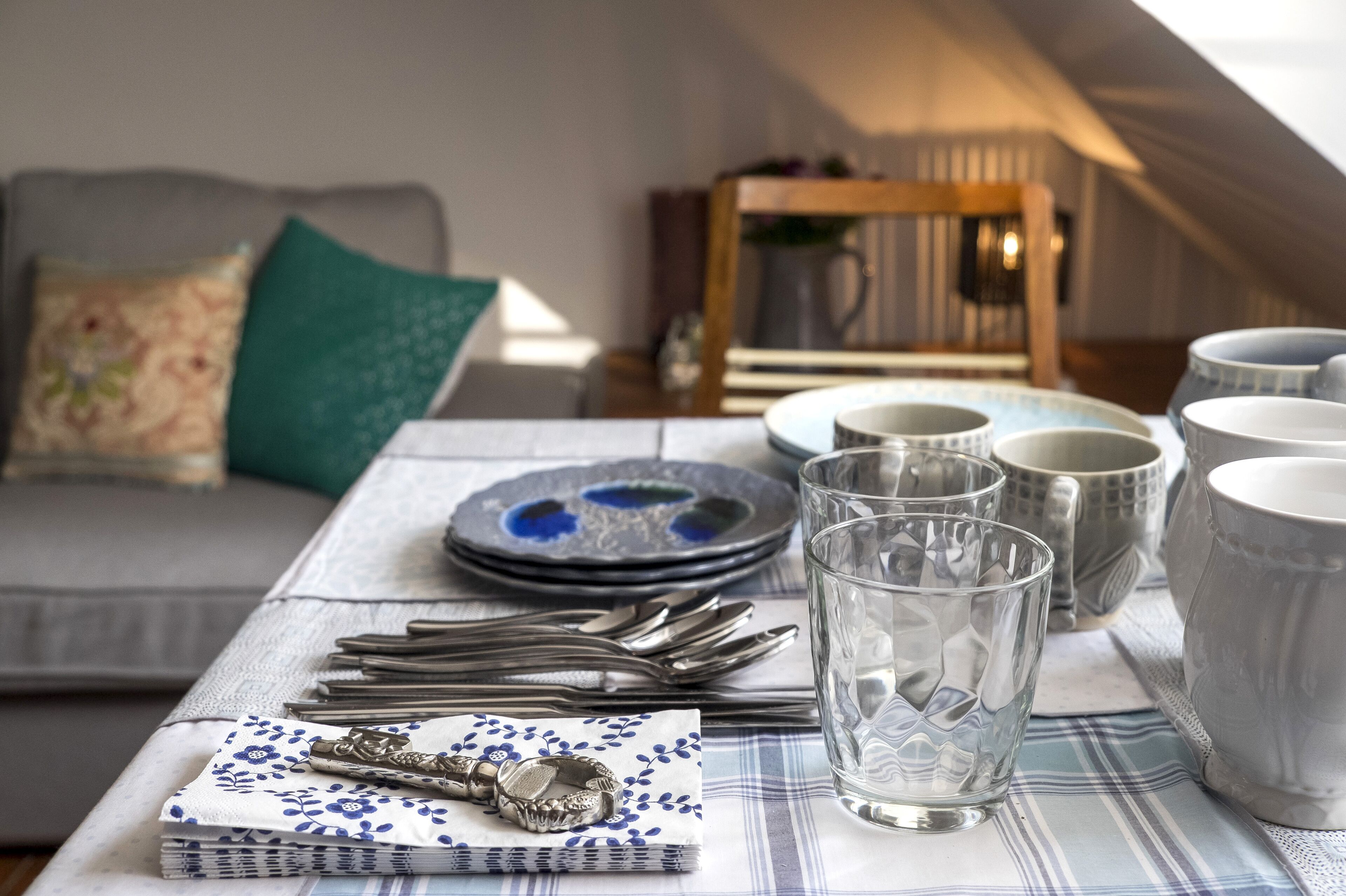 Una mesa de comedor dispuesta con platos, vasos y cubiertos, con un sofá y cojines al fondo.