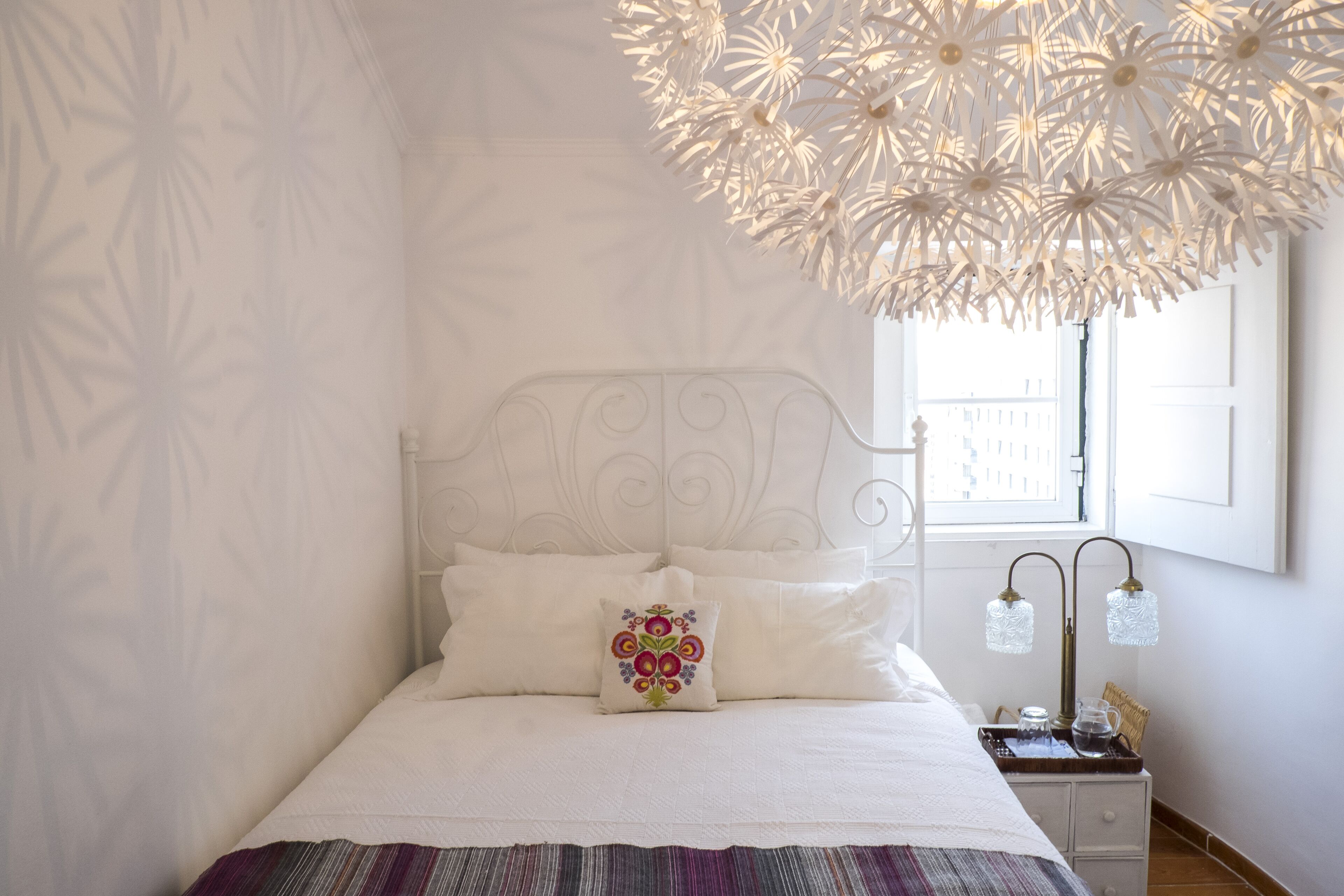Un lit bien fait, agrémenté d’un coussin décoratif, une table de chevet avec une lampe et une fenêtre donnant sur des immeubles.
