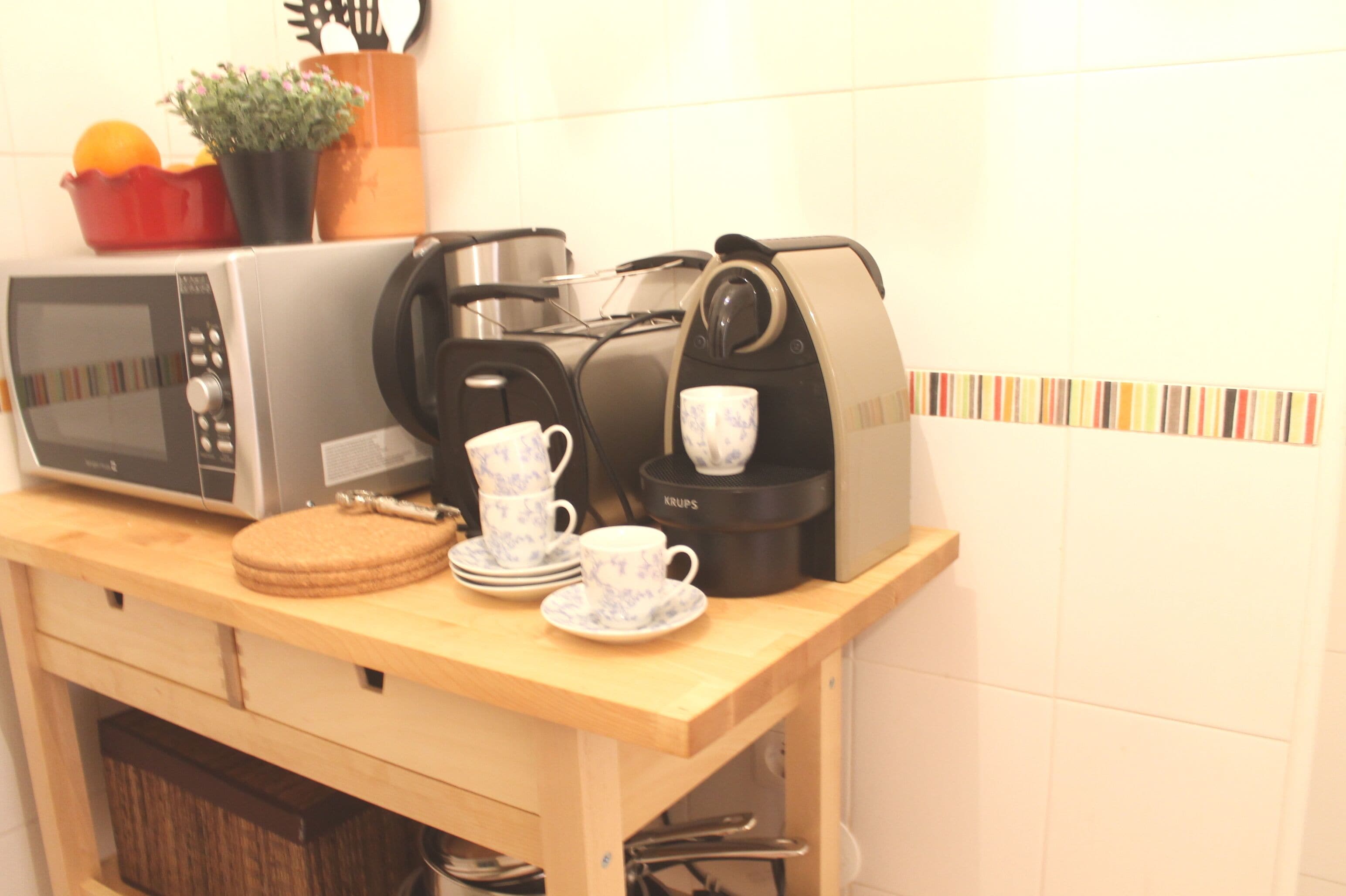 Eine Küche mit Mikrowelle, Kaffeemaschine und einem Holztisch mit Tassen und einer Pflanze.
