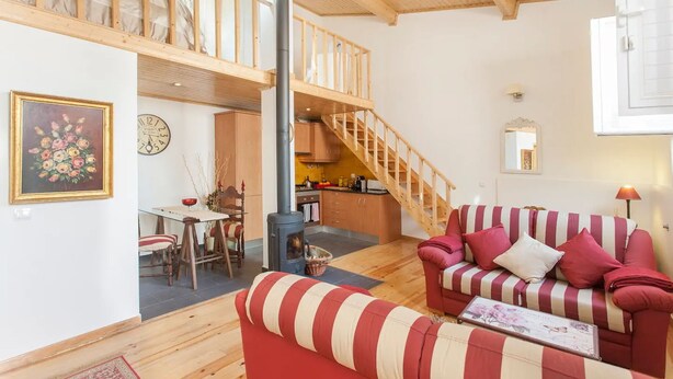 A cozy living room with a red and white striped sofa, a wooden staircase, and a kitchen area.