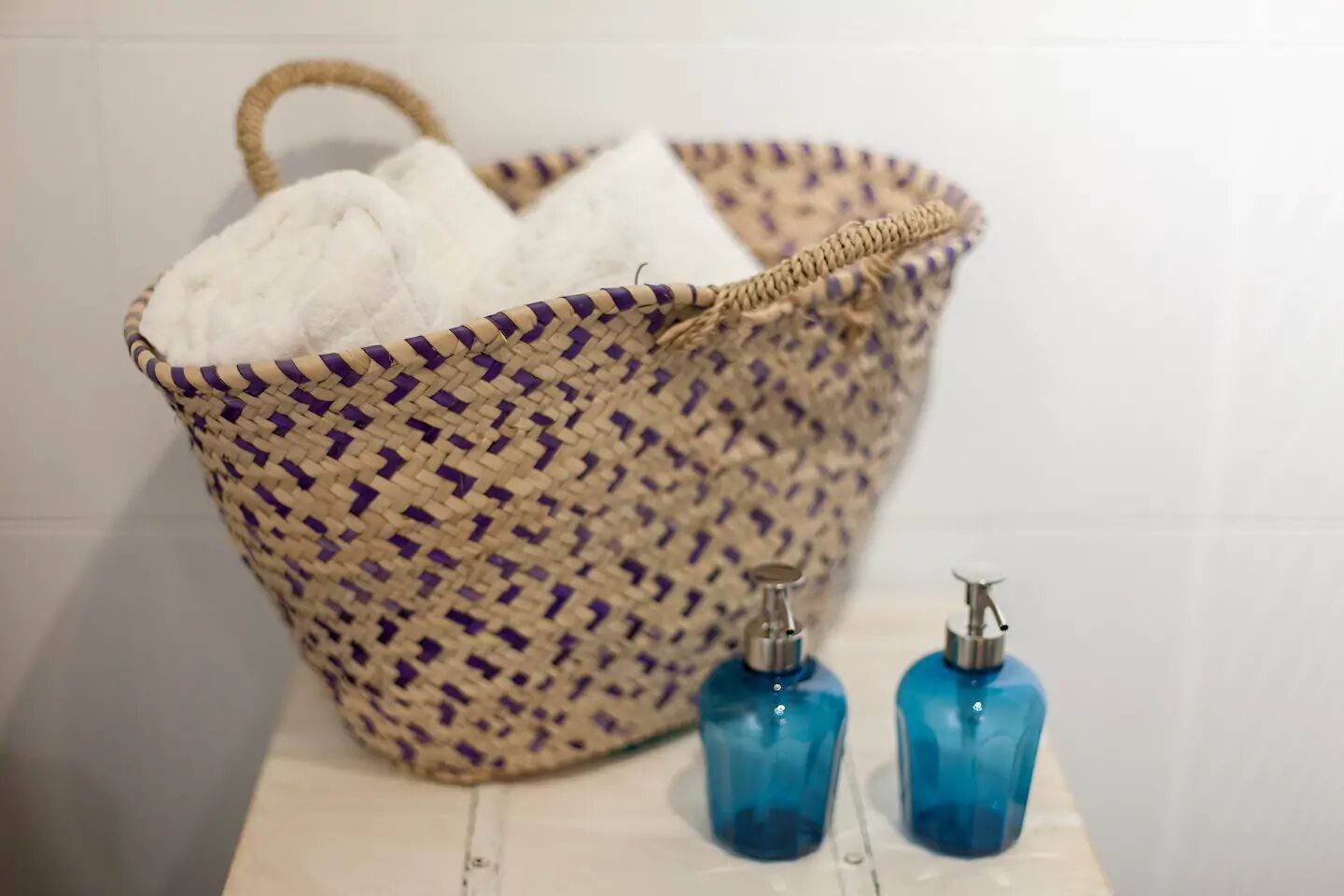 A woven basket with a handle, containing a white towel and two blue bottles on a tiled surface.