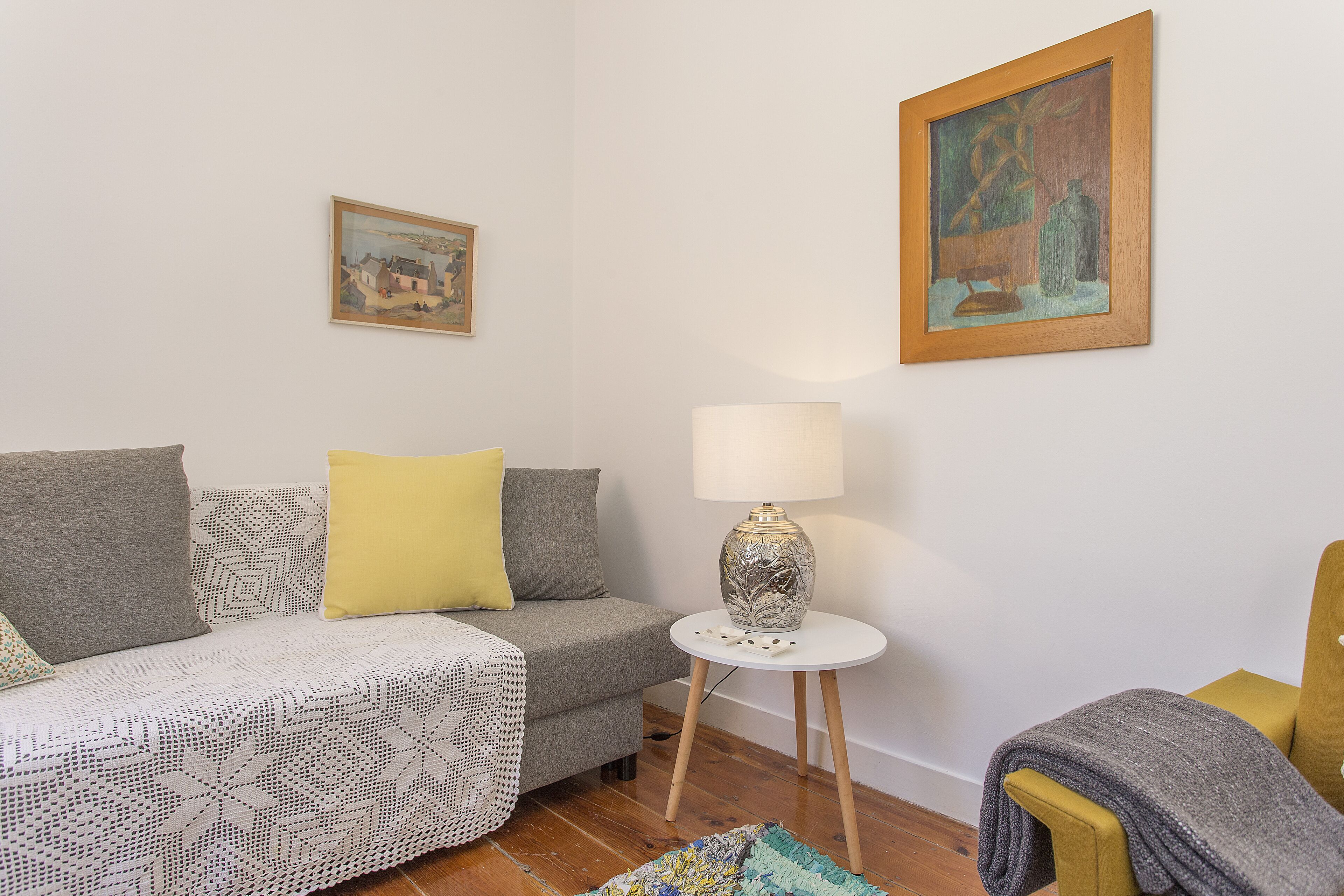 Ein Wohnzimmer mit einer Couch, einem kleinen Tisch mit Lampe, einem Sessel und zwei gerahmten Kunstwerken an der Wand.