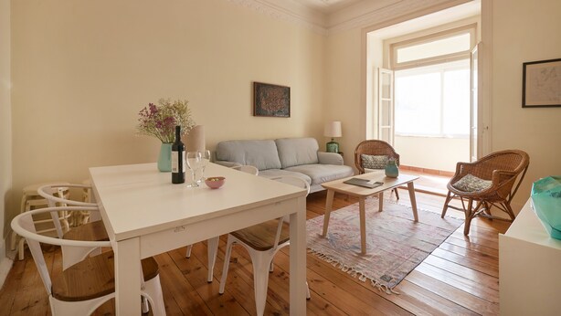 Un salon avec une table blanche, un canapé gris, des fauteuils en osier, un lustre et une grande fenêtre.