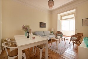 A living room with a white table, a grey sofa, wicker chairs, a chandelier, and a large window. - Renovated Spacious Baixa Apartment, By TimeCooler (Lisbon)