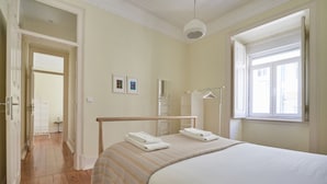 Une chambre à coucher comprenant un lit, une table de chevet, un luminaire suspendu, une fenêtre avec des stores et une porte donnant sur une autre pièce.
