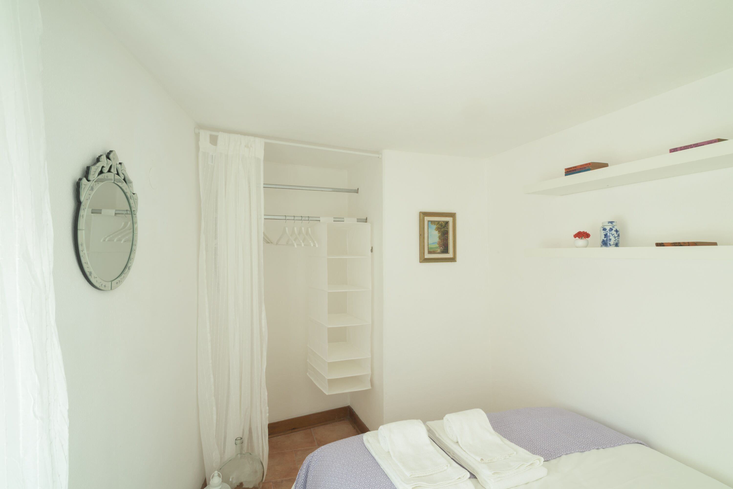 Ein Schlafzimmer mit einem Bett, einem Kleiderschrank, einem Spiegel und einem Regal mit Büchern und einer Vase.