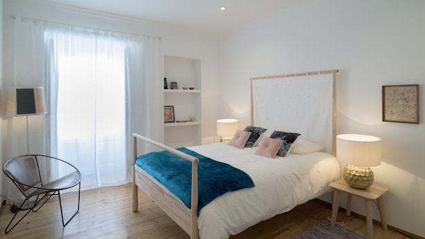 Un dormitorio con cama, mesitas de noche, una silla, una ventana con cortinas y una estantería con objetos.