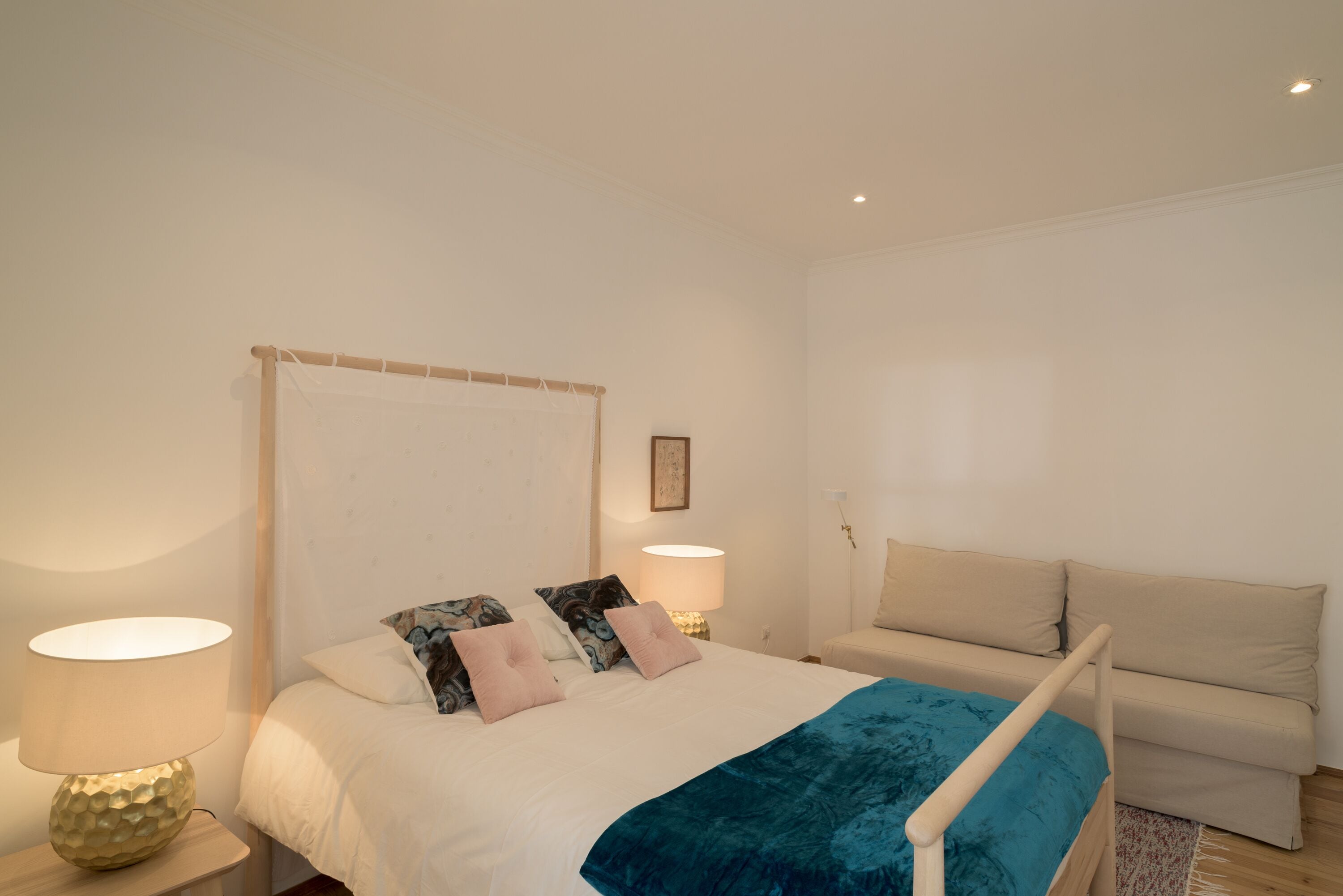 Quarto com cama, luminárias de cabeceira, mesa de cabeceira, sofá e um quadro na parede.