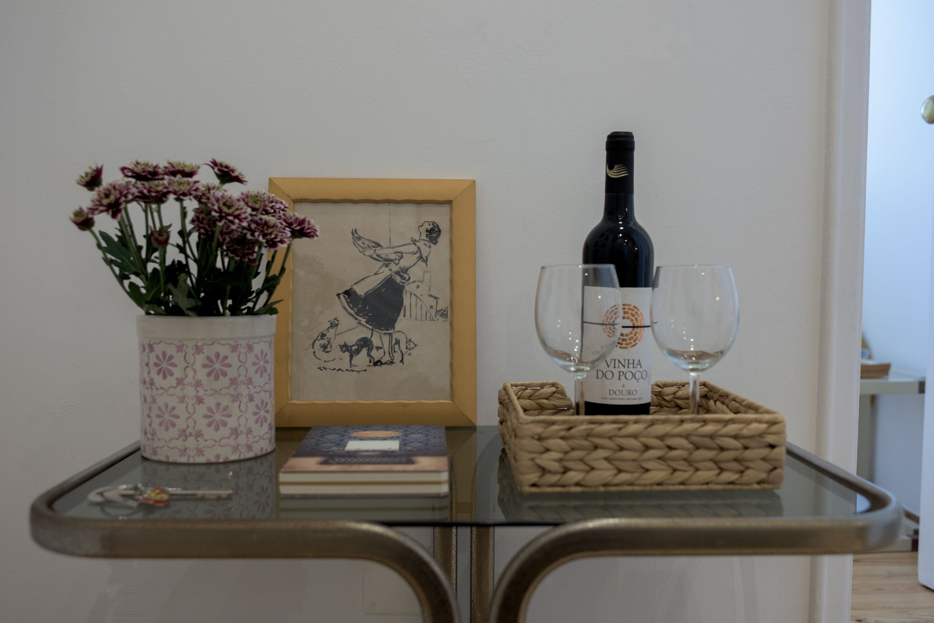 A table with a vase of flowers, a framed picture, a bottle of wine, and two glasses.