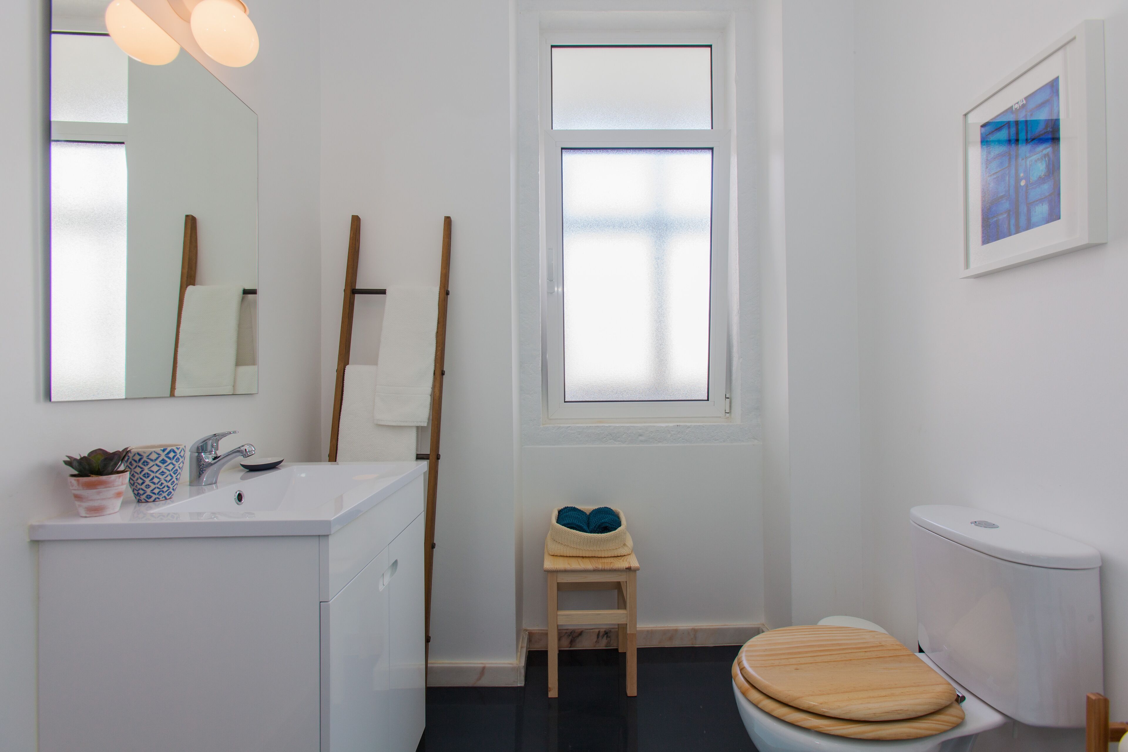 A modern bathroom with a white sink, mirror, and toilet. There is a small stool and a potted plant on the sink counter.
