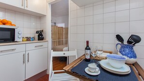 Een kleine keuken met witte kastjes, een magnetron en een koffiezetapparaat. Een eettafel met een placemat, een fles wijn en een schaal is zichtbaar.