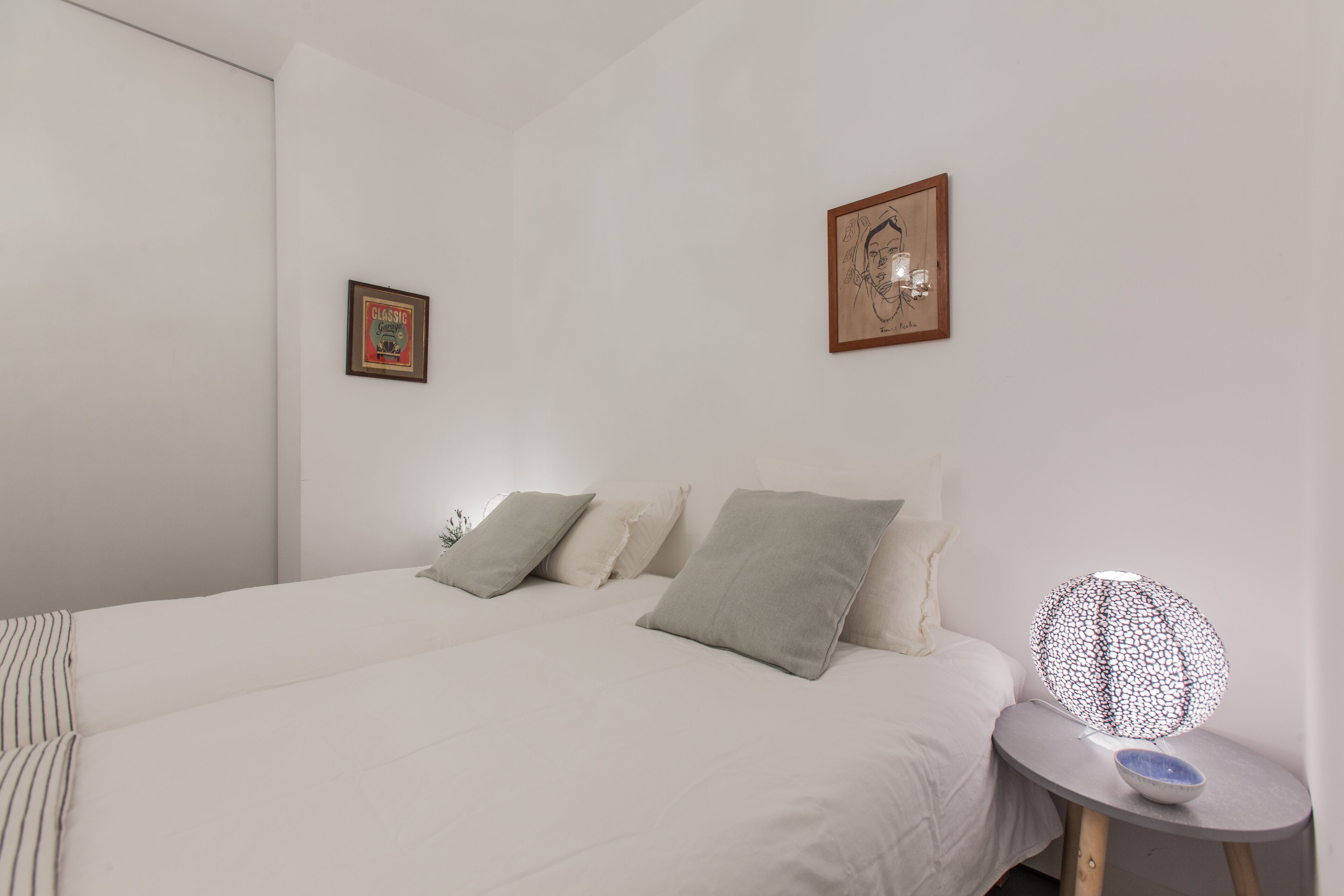 A bedroom with a large bed, two pillows, a bedside table with a lamp, and two framed pictures on the wall.