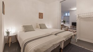 A bedroom with two beds, a bedside table with a plant, a hanging light fixture, and a view of the bathroom through an open door. - Renovated Apartment w/ Private Courtyard, By TimeCooler (Lisbon)