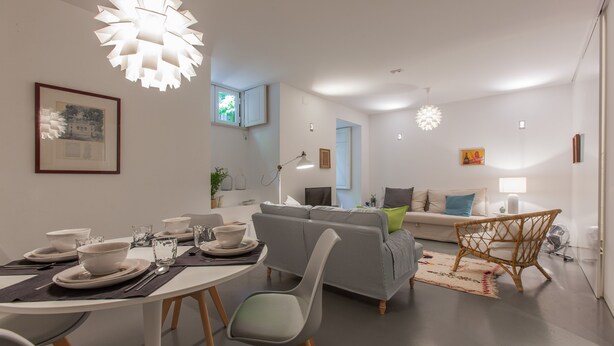 A modern living room with a grey sofa, a dining table set with plates and glasses, and a large white pendant light. - Renovated Apartment w/ Private Courtyard, By TimeCooler (Lisbon)