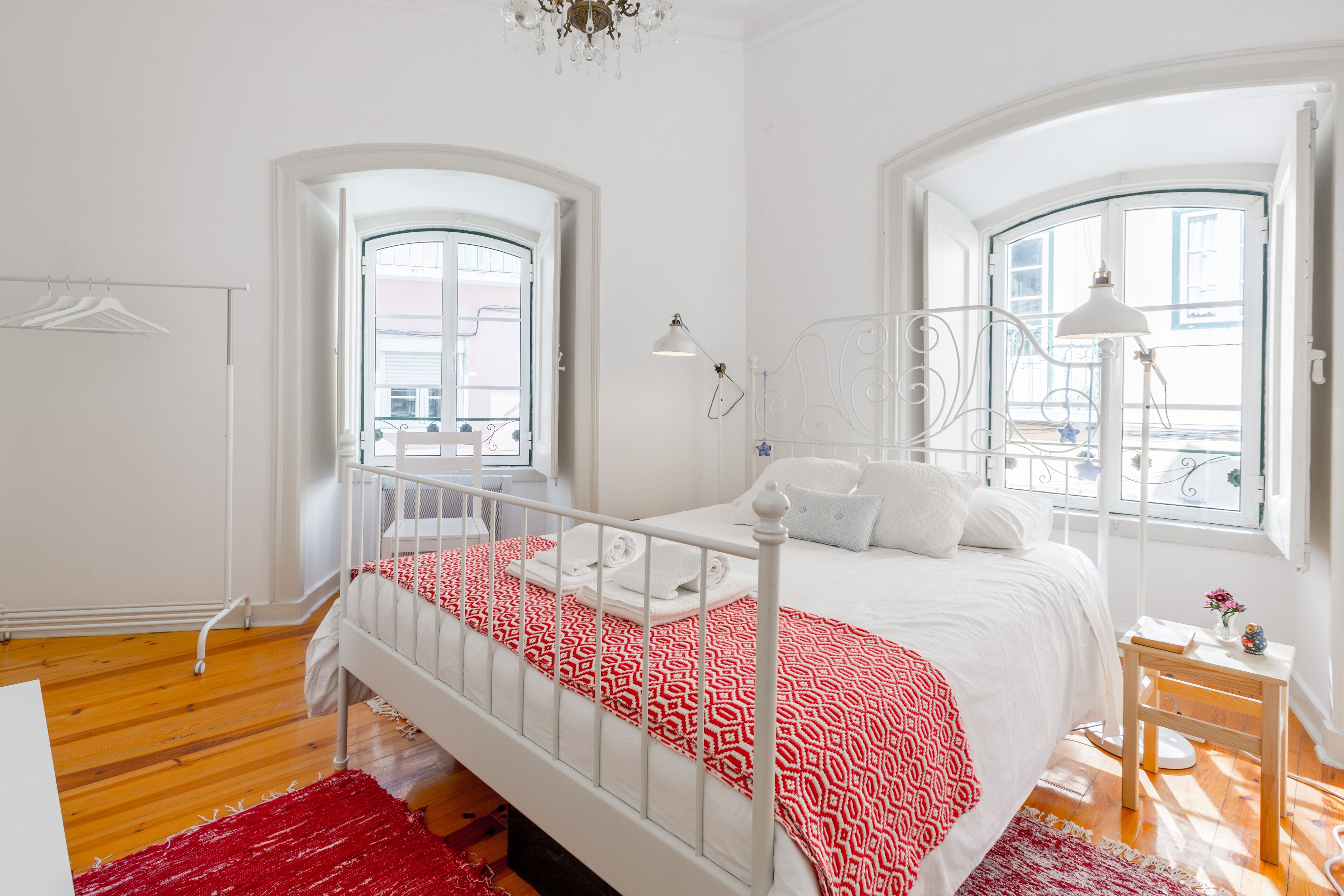 A bedroom with a white bed, red patterned bedspread, and a wooden floor.