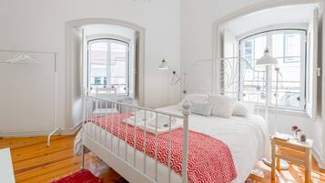 A bedroom with a white bed, red patterned bedspread, and a wooden floor.