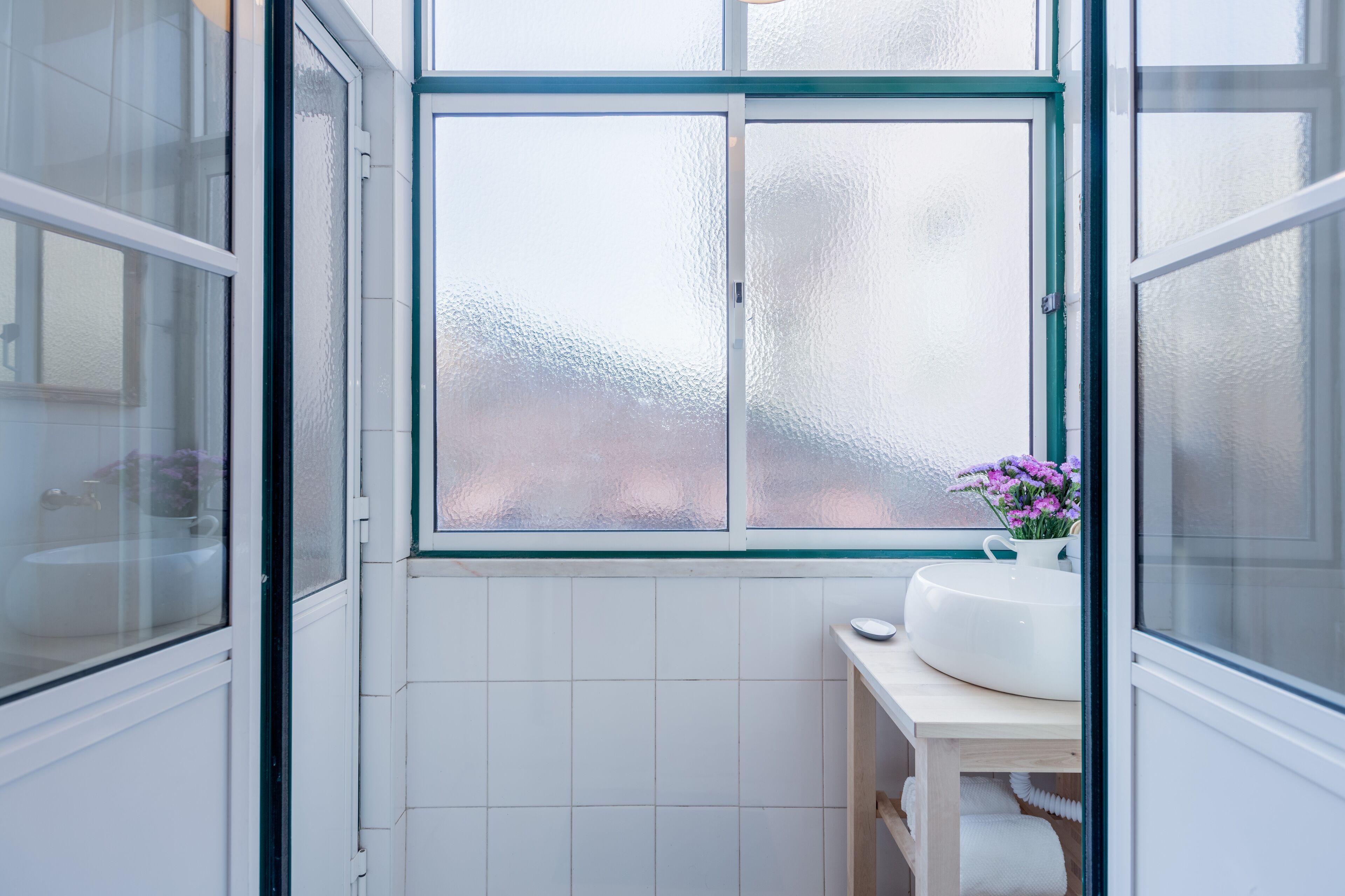 Une salle de bain avec une fenêtre en verre dépoli, un lavabo blanc et une petite table sur laquelle est posé un vase de fleurs.