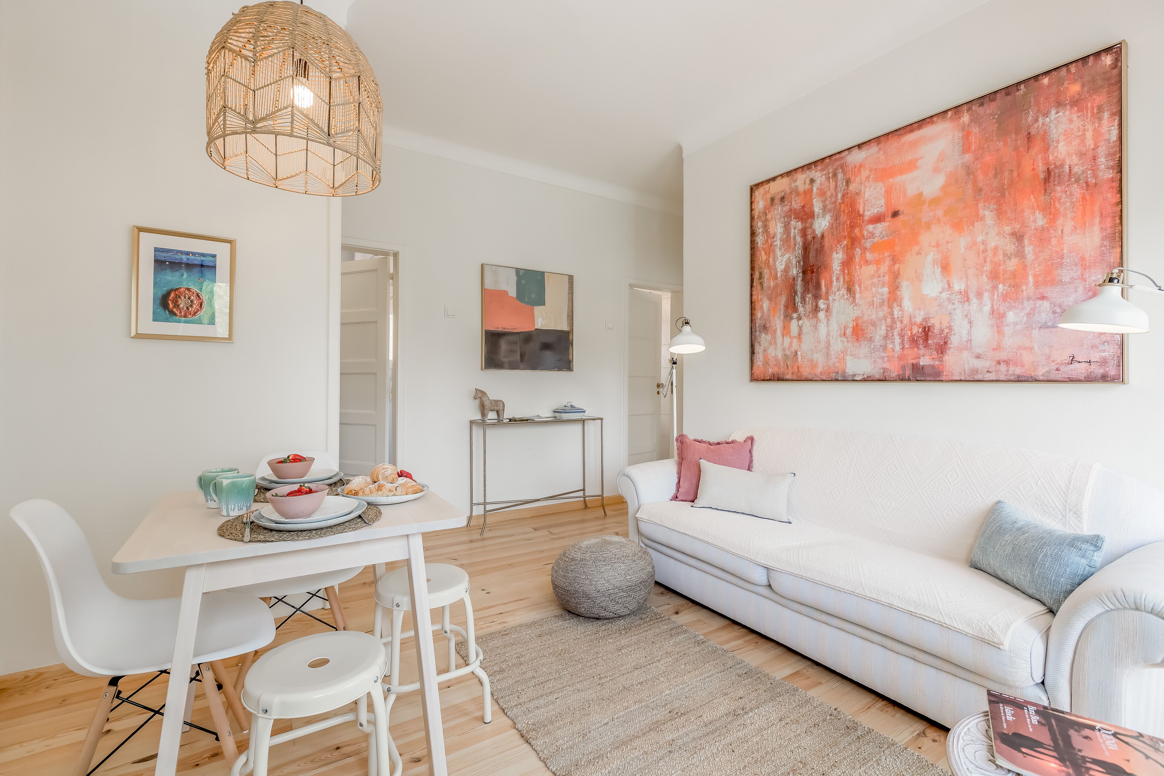 Una sala de estar moderna con un sofá blanco, una mesa de comedor con sillas, un cuadro abstracto de gran tamaño y una lámpara colgante.