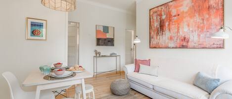 Una sala de estar moderna con un sofá blanco, una mesa de comedor con sillas, un cuadro abstracto de gran tamaño y una lámpara colgante.