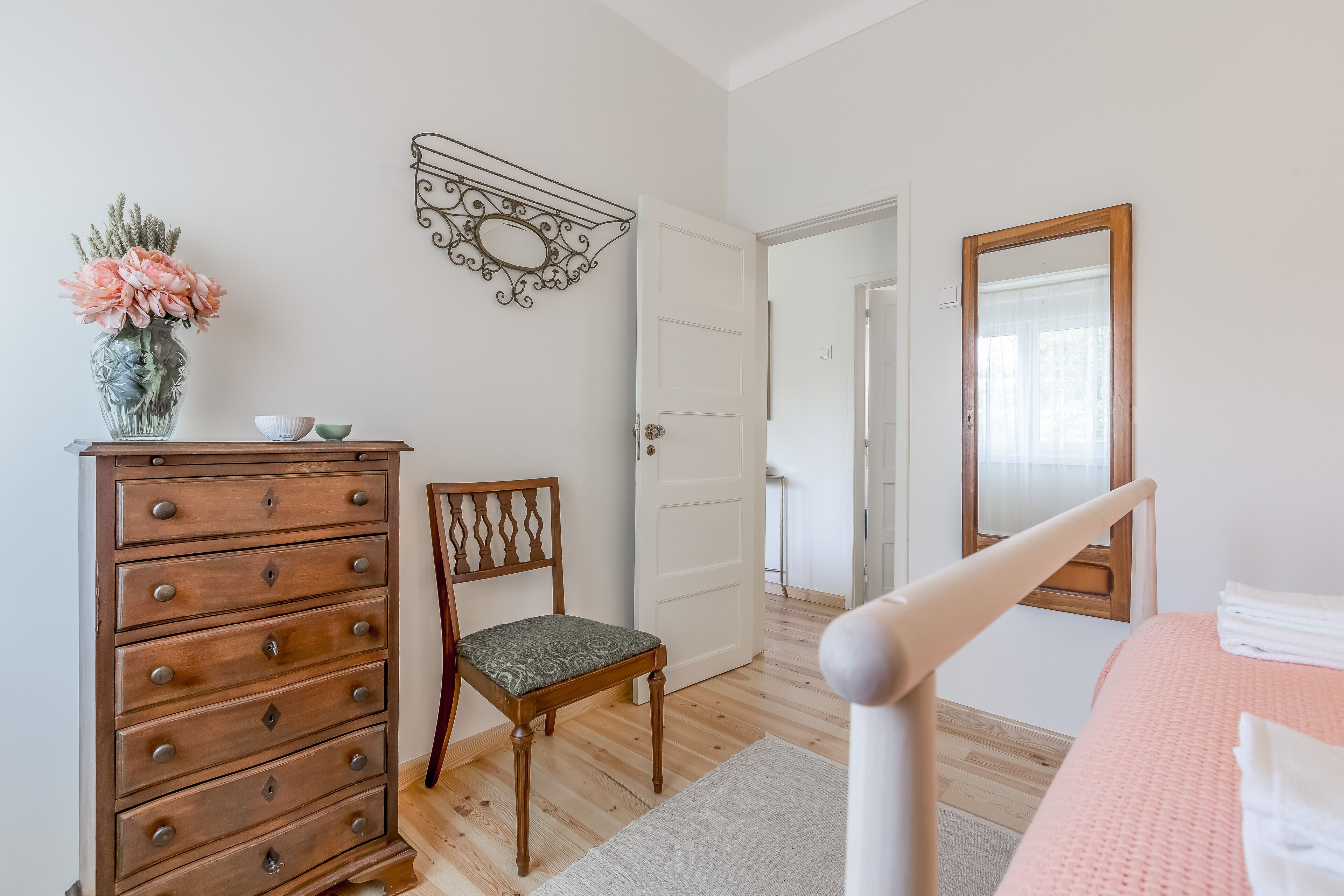 Un dormitorio con un aparador de madera, una silla, una cama con colcha rosa, un espejo y una puerta que da a otra habitación.